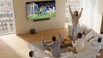 Tres amigos celebran un gol viendo fútbol en una TV grande en su sala de estar moderna con vista a la ciudad. Hay bocadillos y bebidas en la mesa
