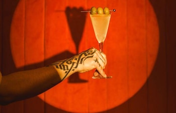 Mano tatuada que sostiene una copa de cóctel con tres aceitunas sobre un fondo circular iluminado en tono naranja