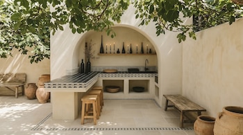 Cozinha externa com paredes creme, bar com azulejos pretos e brancos, cinzeiros, prateleiras com velas e garrafas, cadeiras de madeira, potes de terracota. Folhas verdes superiores.