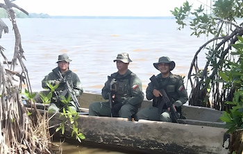 Tres militares uniformados con fusiles y sombreros viajan a bordo de una canoa junto a la orilla de un cuerpo de agua con vegetación