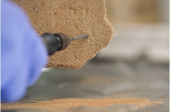 Primer plano de una mano con guante azul que utiliza una herramienta rotatoria en un fragmento de cerámica de color marrón. Polvo fino cae sobre una superficie