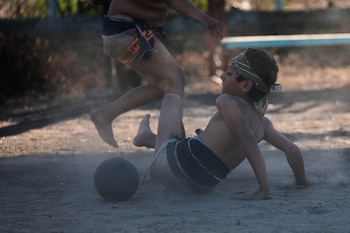 Dos niños descalzos juegan con una pelota oscura sobre tierra polvorienta. Un niño con pañuelo en la cabeza y taparrabos está en el suelo; otro, similarmente vestido, corre detrás