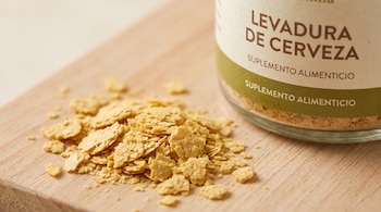 Escamas de levadura de cerveza amarillo dorado sobre madera clara. Parte de un frasco desenfocado con la etiqueta 'Levadura de Cerveza. Suplemento alimenticio'.