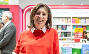 Una mujer senior de cabello castaño sonríe a cámara con un suéter rojo y cuello blanco, de pie frente a estanterías con muchos libros y un cartel de oferta