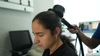 Perfil izquierdo de Raquel, con los ojos cerrados, mientras un técnico aplica un dispositivo de estimulación magnética transcraneal en la parte posterior de su cabeza