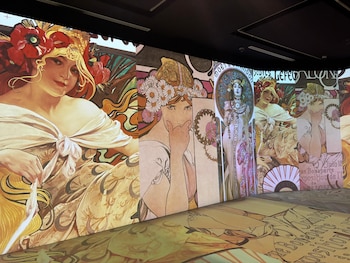 Grandes pantallas iluminadas en una sala oscura proyectan ilustraciones al estilo de Alphonse Mucha con retratos de mujeres estilizadas