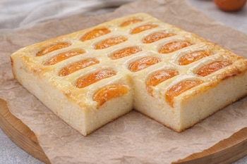 Pastel cuadrado con superficie de crema blanca y mitades de albaricoque. Una porción del pastel está cortada. Se encuentra sobre papel de horno y una tabla de madera.