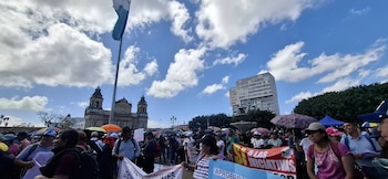 Numerosas personas congregadas en una plaza con una bandera de Guatemala, la fachada de una catedral y un edificio alto. Varias pancartas y sombrillas