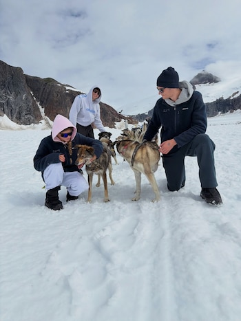 Tres personas y varios perros de trineo en un terreno cubierto de nieve con montañas rocosas al fondo bajo un cielo nublado