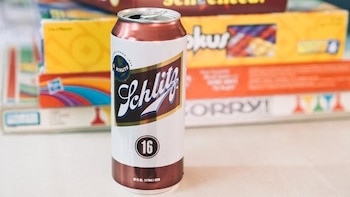 Primer plano de una lata de Cerveza Schlitz abierta, de color blanco y cobre, con el logo negro, frente a cajas de juegos de mesa borrosas