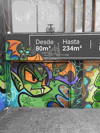 Mural con un personaje verde de orejas puntiagudas y alas sobre un portón, situado bajo un cartel con medidas en metros cuadrados