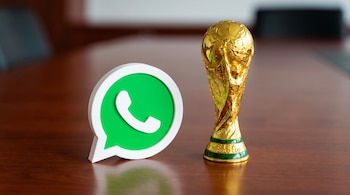 Un logo tridimensional de WhatsApp verde y blanco junto a un trofeo dorado de la Copa Mundial de la FIFA sobre una mesa de madera marrón.