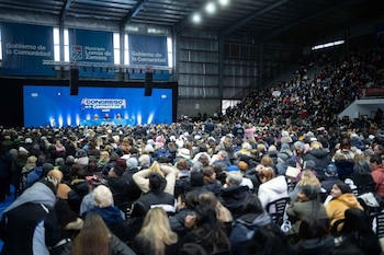 Tres personas saludan desde un escenario con un cartel azul de fondo ante un público que levanta las manos