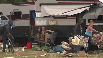 Un hombre en bicicleta, una mujer y otro hombre junto a un remolque gris. Carritos de compras con pertenencias y desechos esparcidos en un campamento