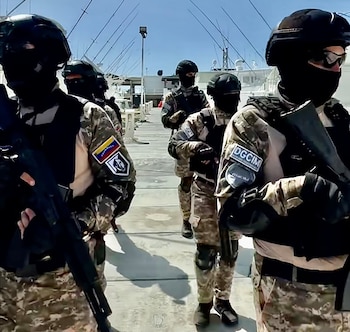 Varios funcionarios de seguridad encapuchados y armados con fusiles, vestidos con uniformes de camuflaje, marchan por un muelle junto a barcos