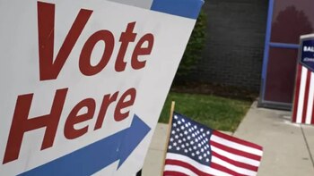 Un cartel blanco con las palabras rojas "Vote Here" y una flecha azul, junto a una pequeña Bandera de los Estados Unidos en el suelo