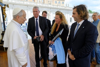 El Papa Francisco, vestido de blanco, conversa con un hombre senior en traje oscuro; detrás, una mujer sostiene un marco y tela celeste