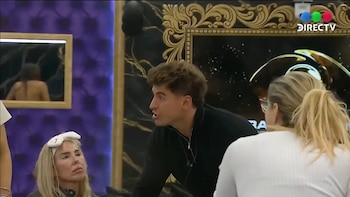 Tres participantes de Gran Hermano en una habitación con decoración ostentosa. Un hombre habla con gesto enfático; dos mujeres lo escuchan atentamente. Una persona se refleja en un espejo