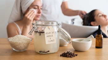 Una mesa de madera con un cuenco de arroz, un frasco de agua de arroz con clavos, clavos secos, un mortero, un frasco gotero y dos mujeres en rutinas de belleza.