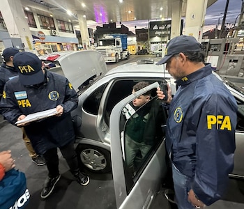 Tres agentes policiales con chaquetas de la PFA inspeccionan a un hombre dentro de un automóvil gris en una estación de servicio