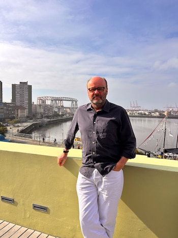 Un hombre con anteojos, camisa oscura y pantalón blanco posa en una terraza amarilla ante un río, varios edificios y un puente de hierro