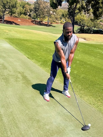Kevin Hart, con camiseta gris sin mangas y pantalones azul marino, se inclina para golpear una bola de golf con un driver en un campo verde y soleado
