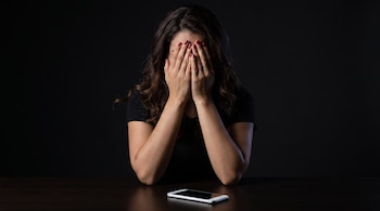 Mujer sentada con las manos sobre su rostro, cabello castaño y uñas pintadas de rojo. Un teléfono celular plateado descansa sobre una mesa oscura.