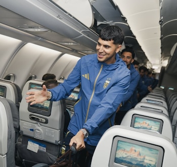 Un futbolista de la Selección Argentina, con chaqueta azul y oro, sonríe y gesticula en el pasillo de un avión. Otros jugadores y pantallas en los asientos