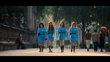 Cuatro mujeres con uniforme azul, cinturones negros y botas caminan en primer plano. Hay árboles y estructuras de piedra en el fondo