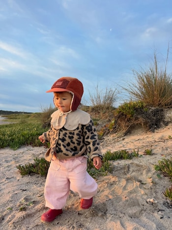 Niño pequeño con gorro marrón, suéter estampado de leopardo, pantalones rosas y botas rojas, caminando sobre arena con vegetación y cielo nublado al fondo