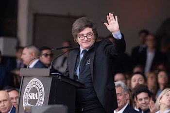 Un hombre con gafas, camisa celeste, corbata negra y saco oscuro saluda con la mano derecha levantada desde un atril con micrófonos