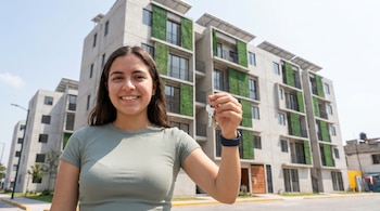 Primer plano de una mujer joven sonriente con cabello oscuro, sosteniendo un manojo de llaves, frente a un moderno edificio de apartamentos con balcones verdes y paneles solares.
