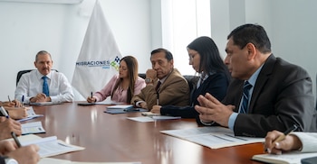 Un grupo de funcionarios se reúne alrededor de una mesa de conferencia con cuadernos y una bandera institucional al fondo