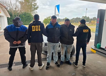Cinco hombres de pie frente a un automóvil en una estación de servicio YPF. Tres llevan chaquetas DDI, uno lleva uniforme YPF y dos llevan abrigos oscuros