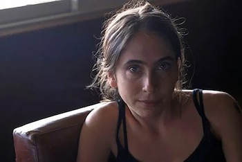 Mujer joven sentada, con cabello oscuro y hebras claras, ojos oscuros y top negro de tirantes, en un interior con fondo oscuro y ventana