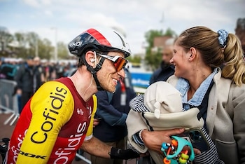 Primer plano de un ciclista sonriente con casco y gafas, mirando a una mujer que sostiene a un bebé en un portabebés. Ambos están sonriendo