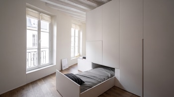 Una habitación con paredes blancas, suelo de madera, grandes ventanas y un armario blanco del cual se despliega una cama.
