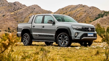 Una pickup Volkswagen Amarok gris estacionada en un campo con pasto seco frente a montañas y cielo nublado. Tiene emblema V6 y patente argentina