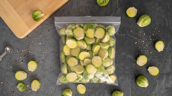 Bolsa de plástico con coles de Bruselas cortadas, variadas coles sueltas, una tabla de madera y una cuchara medidora sobre fondo oscuro.