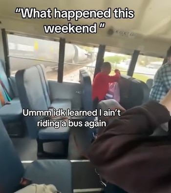 Vista desde el interior de un autobús escolar con asientos azules, mostrando un tren de carga blanco y gris pasando muy cerca de la ventana, con personas a bordo del autobús
