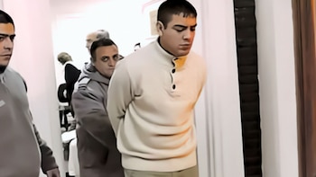 Un hombre joven con corte de pelo corto, vistiendo un suéter crema con cuello abotonado, es escoltado por otro hombre mientras mira hacia abajo