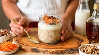 Frasco de vidrio con avena, semillas, yogur, zanahoria y nueces, rodeado por cuencos con nueces, zanahoria rallada, una cuchara y dos botellas.