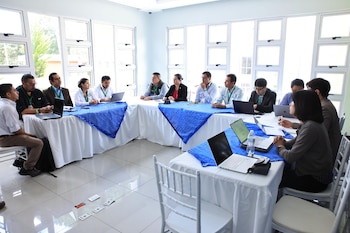 Quince personas sentadas alrededor de una mesa en forma de U con computadoras portátiles y botellas de agua. Hay ventanas grandes detrás. Objetos blancos y naranjas en el suelo