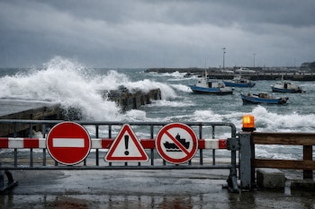 Barrera metálica con tres señales de advertencia y luz naranja frente a un muelle. Olas grandes rompen contra el espigón, con barcos pequeños en el mar agitado y cielo nublado.