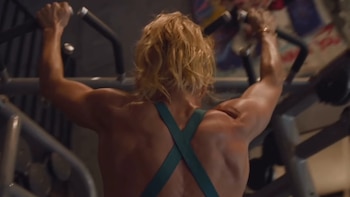 Vista trasera de una persona con cabello rubio y musculatura marcada en brazos y espalda, sosteniendo barras de un equipo de ejercicio en un gimnasio