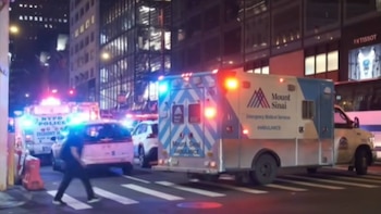 Ambulancia Mount Sinai y coche de policía NYPD con luces de emergencia encendidas en una calle urbana concurrida, un peatón cruza un paso de cebra