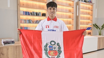 Un joven sonríe y sostiene una bandera de Perú con el escudo nacional. Viste una camiseta blanca con detalles rojos y logotipos. Al fondo hay estanterías con libros