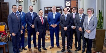 Ocho hombres en traje y corbata y una mujer en chaqueta y pantalón posan para una foto grupal en un interior con piso de mosaico y plantas