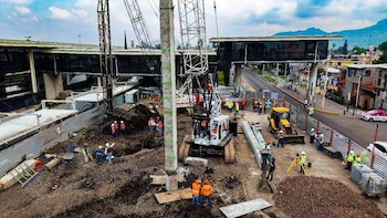 Vista aérea de una obra con maquinaria pesada, excavadoras, grúas y trabajadores con chalecos en una zona urbana junto a un puente peatonal