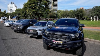 Vista frontal y lateral de varios vehículos decomisados, incluyendo SUVs y camionetas, estacionados en fila sobre asfalto frente a un parque con árboles y edificios